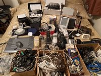 Large assortment of electronic items displayed on a bed including iPod, recorders, Kindle, Wacom tablet, speakers, phones, and many cables in boxes