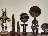 Wide view showing entire lot of six cultural sculptures including Native American Kachina doll with feathers, two Canadian totem poles, and three African wooden sculptures including one large with circular head.