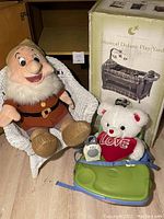 Photo of the lot showing the Disney Doc plush in the white wicker chair, the boxed Eddie Bauer Musical Deluxe Play Yard, the Fisher Price baby chair tray, white teddy bear with red heart, and Sony baby monitor.