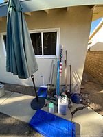 Large assortment of outdoor backyard items arranged near a house wall including umbrellas, shovels, broom, fertilizer, and flag in packaging.