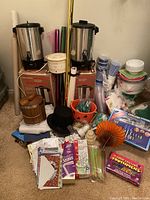 Wide view of the lot including two stainless steel automatic coffee brewers, Rival crock-pot slow cooker, wood ice bucket, various party supplies such as plastic cutlery, cups, plates, gift bags, and holiday decorations.