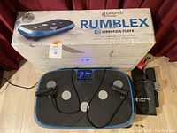 Vibration plate on floor with box and accessories (handles, bands, and storage bag) visible