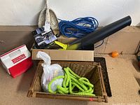 Photo showing a red and white Royalite cooler, woven basket holding a green expandable hose, blue braided hose coiled, a cardboard box containing Ryobi blower and Die Hard battery charger box, black longer plastic tube, and some orange practice golf balls on floor.