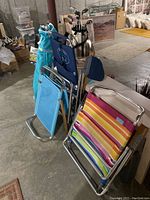 Three folded beach chairs grouped together in basement setting with concrete floor. Includes Tommy Bahama blue recliner, multicolor striped Rio chair, and blue shade recliner with bag.