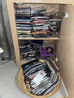 Photo showing a wooden shelf filled with stacks of compact discs in cases, along with additional CDs in a basket below. Cases are a mix of jewel cases with album art visible on some.
