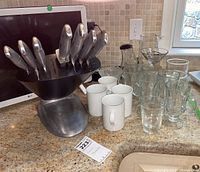 Wide view of the stainless steel knife block with seven knives, four white mugs, and various clear glasses including tumblers and a pitcher on the countertop.
