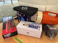 Four items on the floor near a couch: boxed flat iron, boxed projection clock, steam inhaler, and orange flashlight.