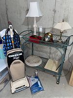 Full view of the vacuum cleaner, lamps, clock, and other items on and around a glass and metal baker's rack in a basement.