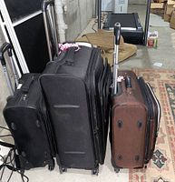 Three soft-sided luggage pieces standing upright showing varying sizes and colors (two black, one brown) with wheels and handles.