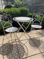 Full view of the white metal bistro set with two folding chairs and a round table placed outside on a patio.