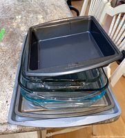 Stack of multiple baking pans including Wilton black non-stick square pans, clear glass dishes, and metal baking trays