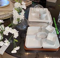 Full view of snack set with multiple white ceramic dishes arranged on a dark wooden tray, a white ceramic mug on top, and clear glass vase with artificial white flowers next to them on a glass table.