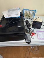 HP OfficeJet 4630 printer with manual and cords, stapler, hole punch, scissors, Sony Dream Machine clock radio, blue pouch with small bottles and other items on white table.