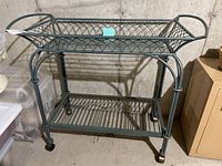 Full view of the metal rolling cart with two wire shelves and four wheels on a concrete floor.