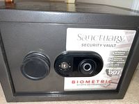 Close-up photo of the front face of the Sanctuary Security Vault showing biometric fingerprint scanner, round dial knob, keyhole without key, and branding label detailing features like interior lighting and tamper indicator.
