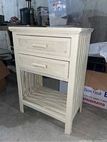 Front view of the wooden side table with two drawers and slatted bottom shelf, showing off-white paint and scratches.