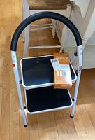 Front view of white metal two-step stool with black steps and curved padded handle, doorbell box placed on top step.
