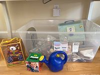 Overview of lot showing all items including teddy bear toy, Plus-Plus block set, blue watering can, baby briefcase with baby book, and plastic bin with candles and holders.
