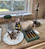 Photo showing ceramic bowls labeled SWEET and SYLVIA, plate with decorative utensils, glass container with green lid, decorative oil bottles, and box of ceramic spreaders with floral designs.