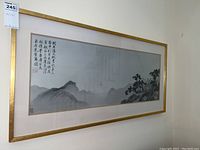 Full view of the framed Asian themed wall art hung on a wall showing gold frame, landscape, and calligraphy.