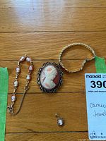Top-down photo of the four pieces laid on a wood surface, showing cameo bracelet, large cameo brooch, small cameo pendant, and gold-tone bracelet together with MaxSold lot tag