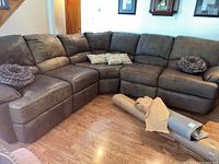Full view of L-shaped brown leather-like 5-piece sectional couch with accent pillows and rolled-up carpet on floor in front.