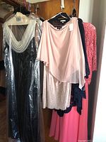 Photo of dresses on hangers showing a black dress with white lace collar, a blush pink chiffon dress with embellishments, a bright pink lace dress, and navy lace shawl and robe.