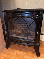 Full view of black Duraflame electric fireplace with curved glass front showing decorative metal cross and faux wood logs inside.