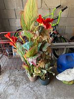 Two tall faux floral plants with vibrant leaves and red flowers in plastic pots in front of a metal cage and a black bicycle