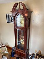 Full frontal view of the grandfather clock showing the wooden ornate top, clock face behind a glass door, and the lower glass panel revealing pendulum and weights.