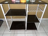 Front and side view of metal island table showing dark wood composite shelves and white metal frame with wire rack on side