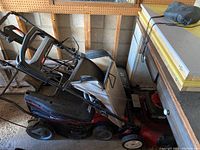 Side view of both lawnmowers stacked in garage, showing handles, wheels, and cloth grass catcher on Toro mower.