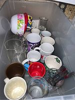 Top view of assorted mugs, glasses, floral bowl, glass carafe, and forks grouped in plastic bin.