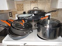 View of all cookware pieces stacked together showing orange-handled Rachel Ray pots and pans, Farberware stockpot with lid, and Metro BBQ Tote in background.