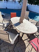 Photo of outdoor oval stone-like topped table with four chairs and central umbrella covered with beige protective sleeve, on brick patio surface.