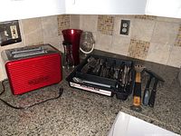Photo of red Sensio Bella toaster, black plastic utensil holder filled with various metal utensils, red vase, glass jar, kitchen hammer and scraper on kitchen counter.