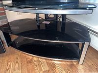 Front angle of black glass and metal TV stand with lower shelf indoors on wood floor.