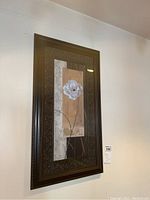 First framed floral wall art piece showing a white flower on a beige and off-white background, in an ornate dark frame