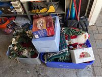Two large plastic totes filled with assorted Christmas decorations including box labeled ceramic light-up tree, wreath, Santa figure, rugs, candle holders, and other holiday decor.