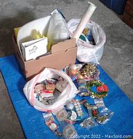 Photo showing large cardboard box, multiple plastic bags filled with craft supplies including ornaments, ribbons, and other decorative items arranged on a blue surface.