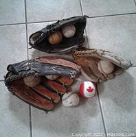 Photo showing the 3 baseball gloves with 7 baseballs, all placed on tiled floor. Gloves include two black Wilson gloves and a tan glove; baseballs include a Canadian ball and several used balls.
