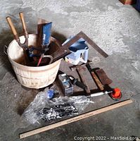 Photo showing wooden bushel basket with multiple hand tools and hardware items placed around it on a concrete floor.