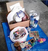 Overview of the large assortment of craft supplies including cardboard boxes, plastic bags, ribbon spools, wicker baskets, and colored eggs on a blue tarp.