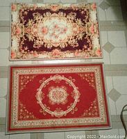 Photo showing two floor mats side by side on tiled floor, one burgundy with floral designs and one red with central medallion.