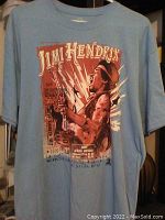 Light blue t-shirt front showing Jimi Hendrix playing guitar with large graphic and text.
