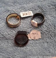 Three men's rings placed on a blue textured fabric, showing one polished silver-tone ring, one black ring with metallic cross design, and one black faceted ring, all tagged with pricing labels.