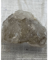 Close-up photo of a raw smoky quartz crystal showing its translucent brown-gray hues and natural uneven crystalline structure on a textured background.