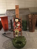 Photo of Bohemian decorative items including vases in red, green, copper colors, a figurine of a lady in traditional attire seated on a wooden chair, bronze candlestick, and metal coatrack.