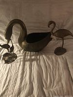 Image of two brass bird sculptures, a large swan-shaped container and a standing flamingo on base, displayed on white fabric background.