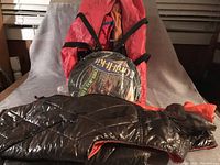 Photo showing a reversible vest in black and red, a small cooking grill wrapped in plastic, and a rolled pop-up tent in a red and blue carrying bag.